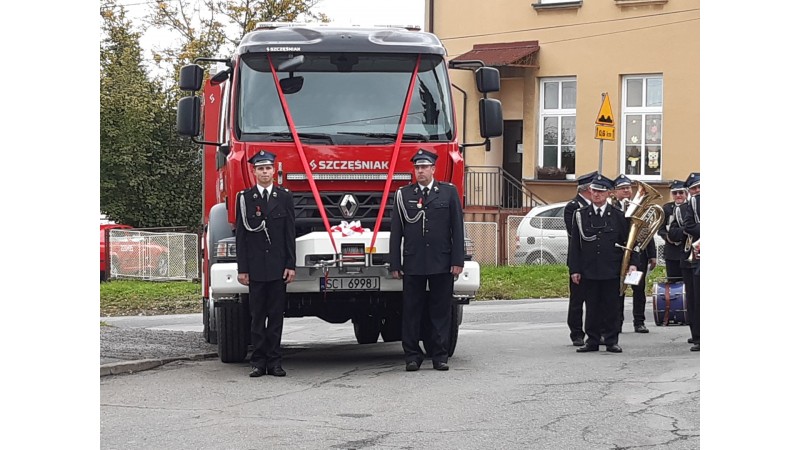 Samochód ratowniczo-gaśniczy GBA Renault z dwoma strażakami po bokach podczas uroczystości przekazania.